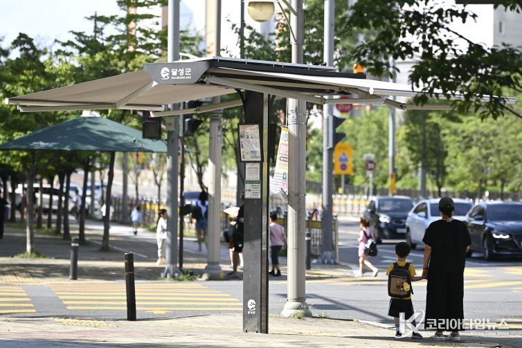 2025년 08월 21일_달성군 폭염 저감 정책(한 모자가 횡단보도 앞에서 신호를 기다리며 설치된 스마트 그늘막 아래에서 햇볕을 피하고 있다.)